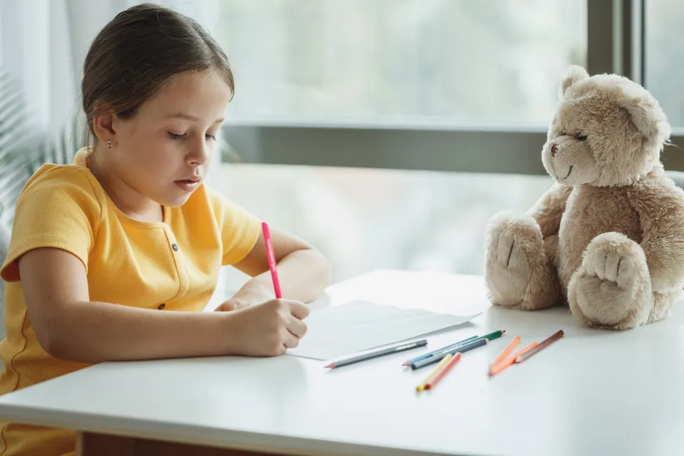 Une enfant fait ses devoirs à une table, crayons de couleur et ours en peluche à ses côtés